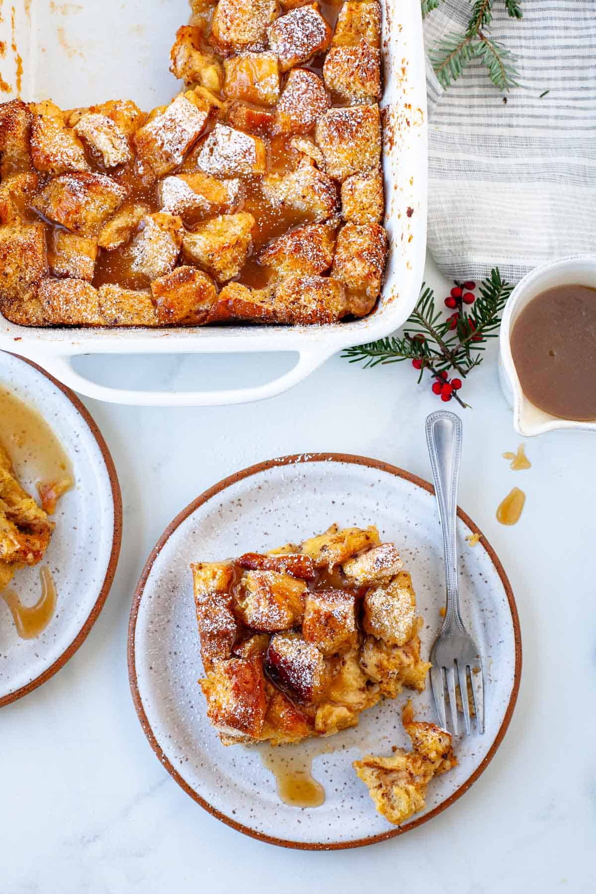 eggnog bread pudding on white speckled ceramic plate with powdered sugar on white background and green Christmas foliage